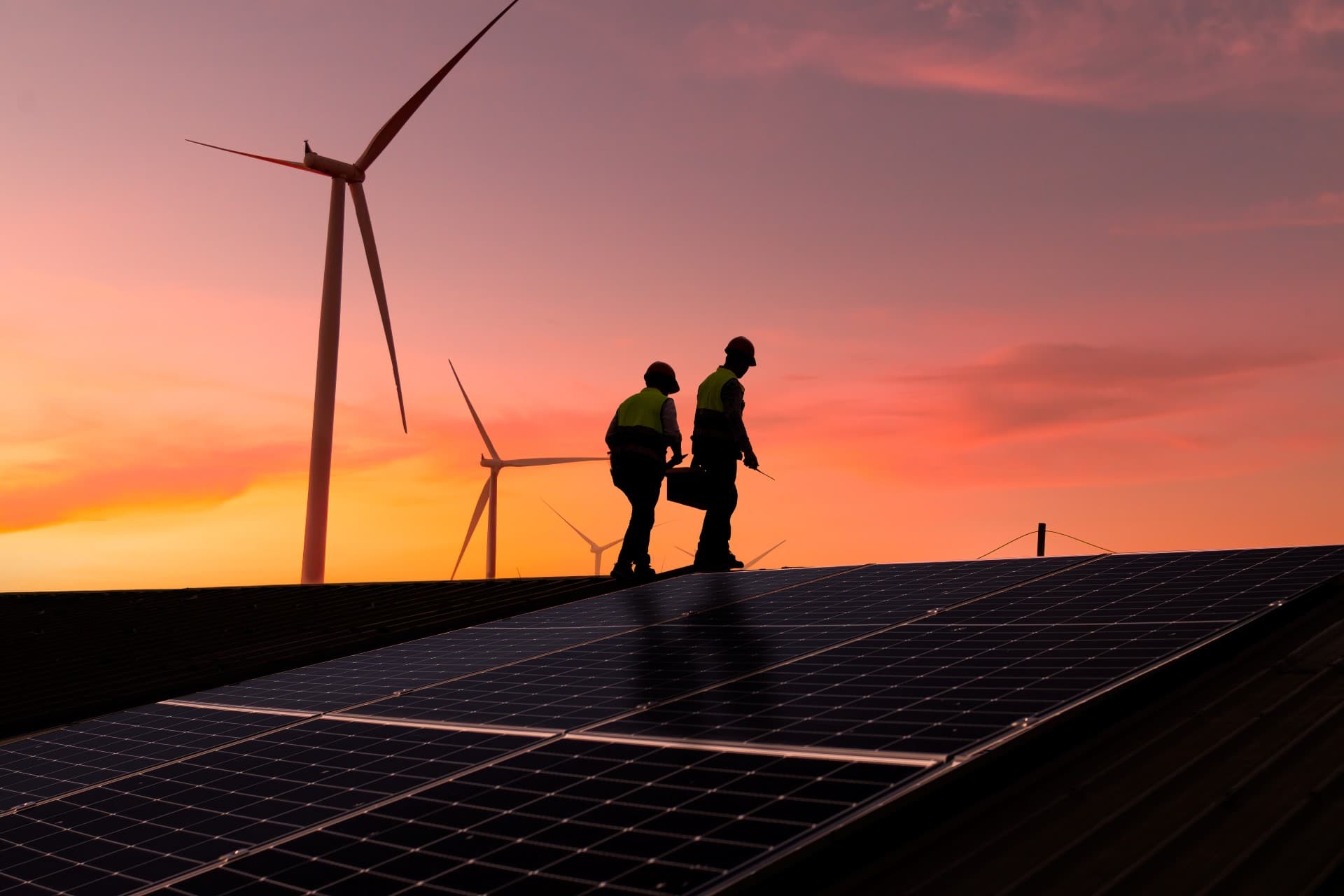 Two engineers inspect a solar panel installation with wind turbines in the background at sunset.