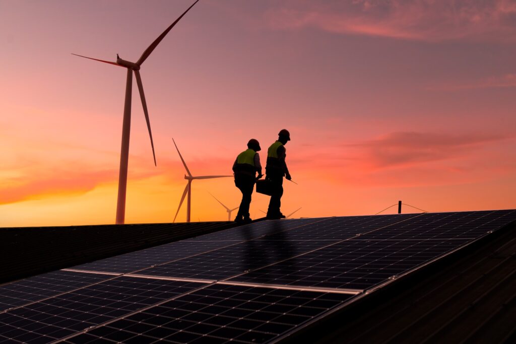 Two engineers inspect a solar panel installation with wind turbines in the background at sunset.