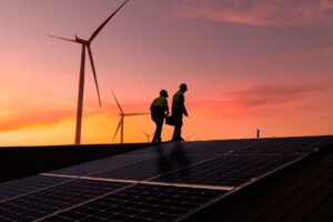 Two engineers inspect a solar panel installation with wind turbines in the background at sunset.