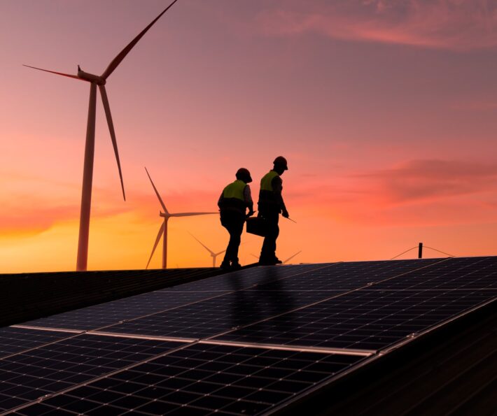 Two engineers inspect a solar panel installation with wind turbines in the background at sunset.